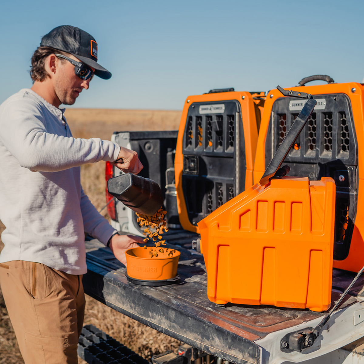 A man wearing sunglasses and a hat pours dog food from a black container into the GUNNER DOG BOWL - BLAZE ORANGE on a truck tailgate, with limited-edition Blaze Orange pet carriers from GUNNER and a clear blue sky in the background.