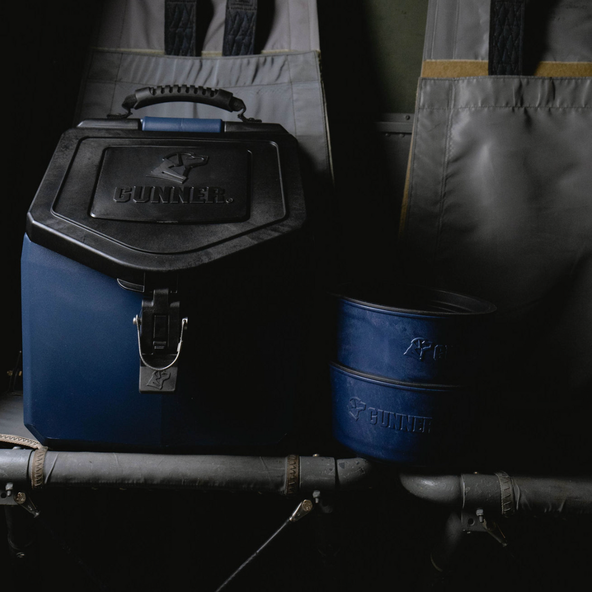 A limited-edition GUNNER DOG BOWL - NAVY sits on a metal bench next to two matching blue containers, with gray padded backrests and metal framework in the background—part of the exclusive GUNNER collection.