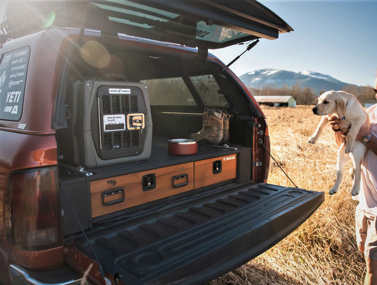 A pickup truck with its tailgate open reveals organized outdoor gear, a portable fridge, boots, and a dog bowl. A person holds a yellow Labrador puppy—future hunting dog—next to the truck in a field with mountains in the background.