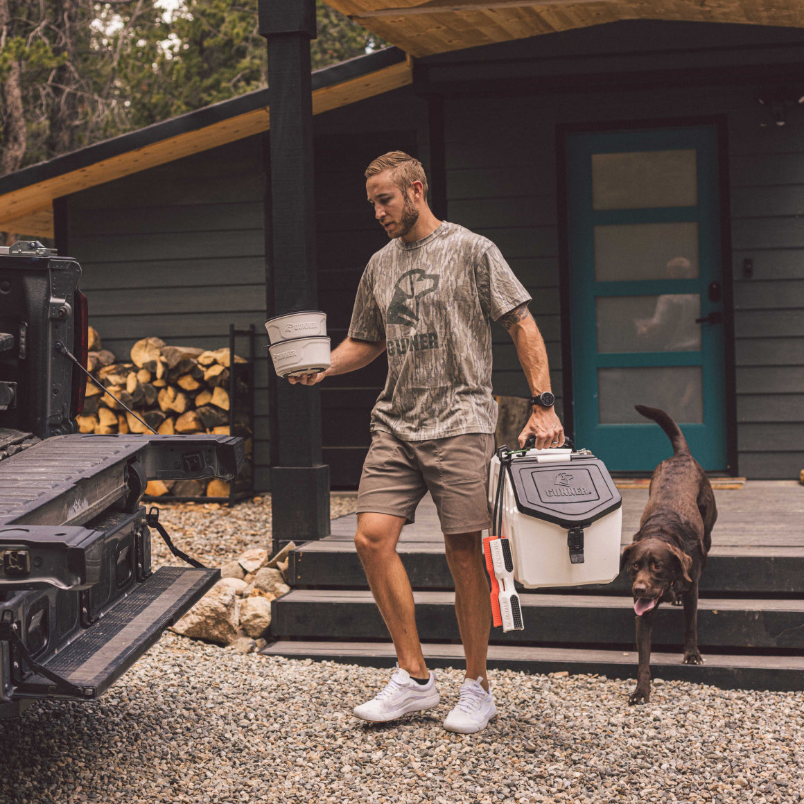 A man in a GUNNER BOTTOMLAND TEE carries a cooler and bowl to a black pickup near the cabin, followed by his black dog on the gravel path. Stacked firewood rests beside the porch under Bottomland trees.