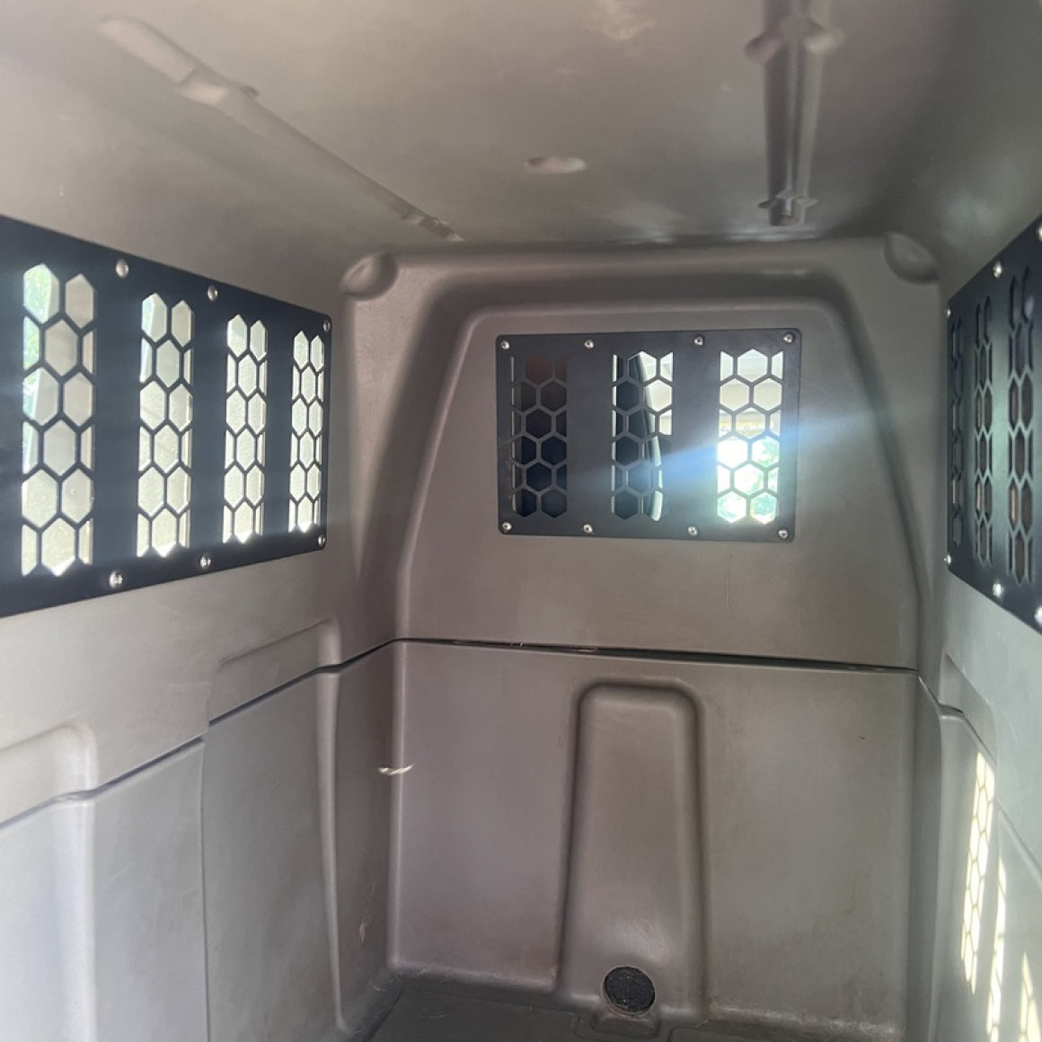 Interior of a metal vehicle compartment with hexagon-patterned vents, likely a transport van. Light filters through vented windows—ideal for securing GUNNER kennels with the CHEW KIT for added dog kennel protection.