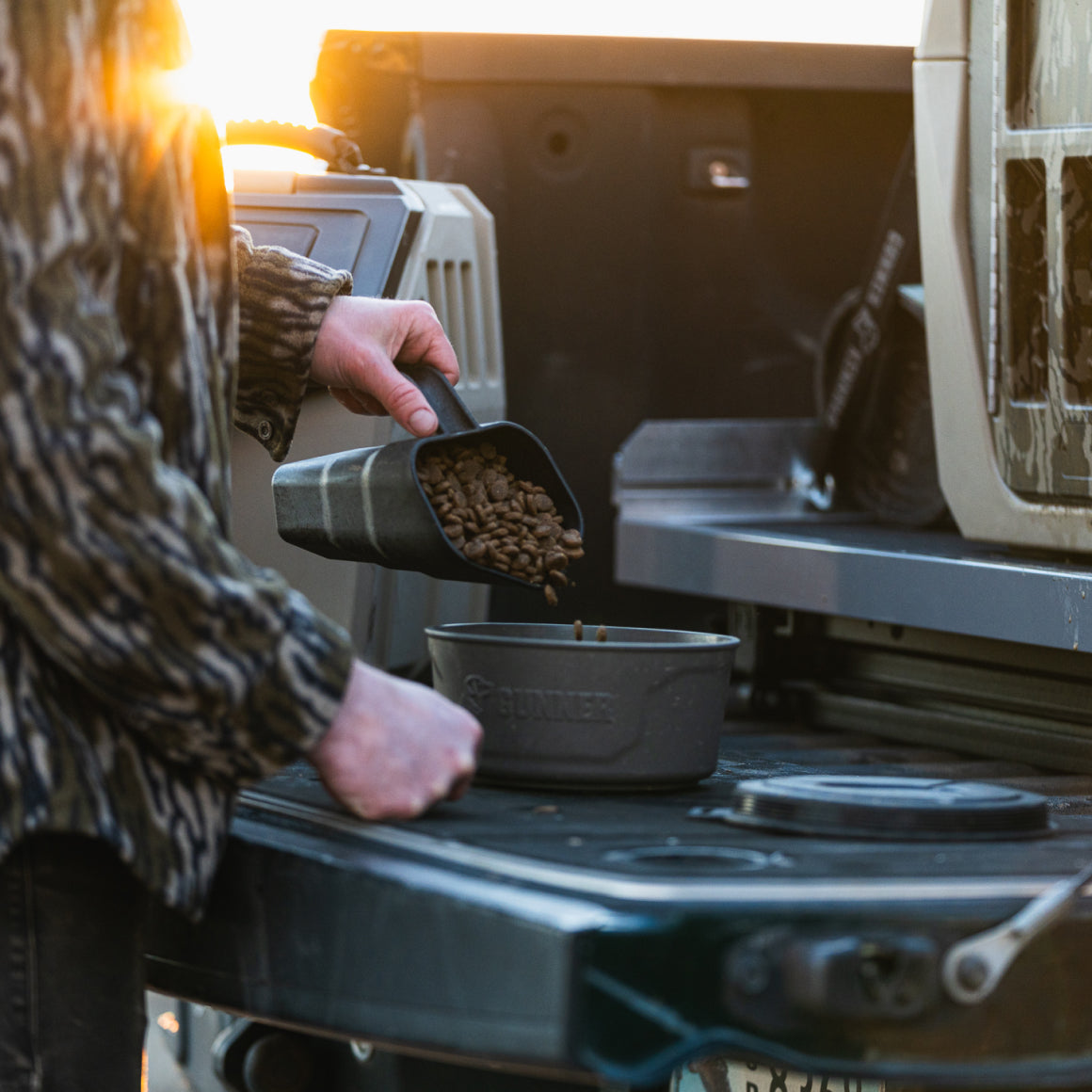 A person uses the GUNNER FOOD SCOOP to pour dry pet food from a container into a bowl on an open vehicle tailgate, with sunlight in the background.