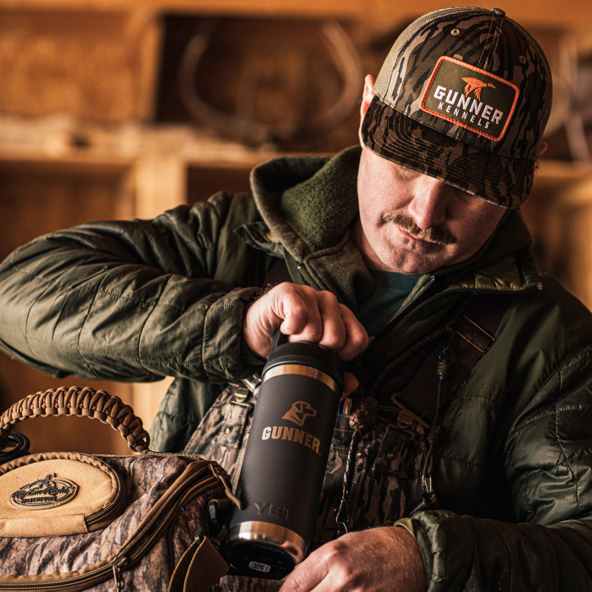 A man in Mossy Oak camo gear and a GUNNER BOTTOMLAND MALLARD TRUCKER hat holds a black GUNNER-branded Yeti bottle, standing indoors with a camouflage bag over his shoulder as he looks down at the bottle.