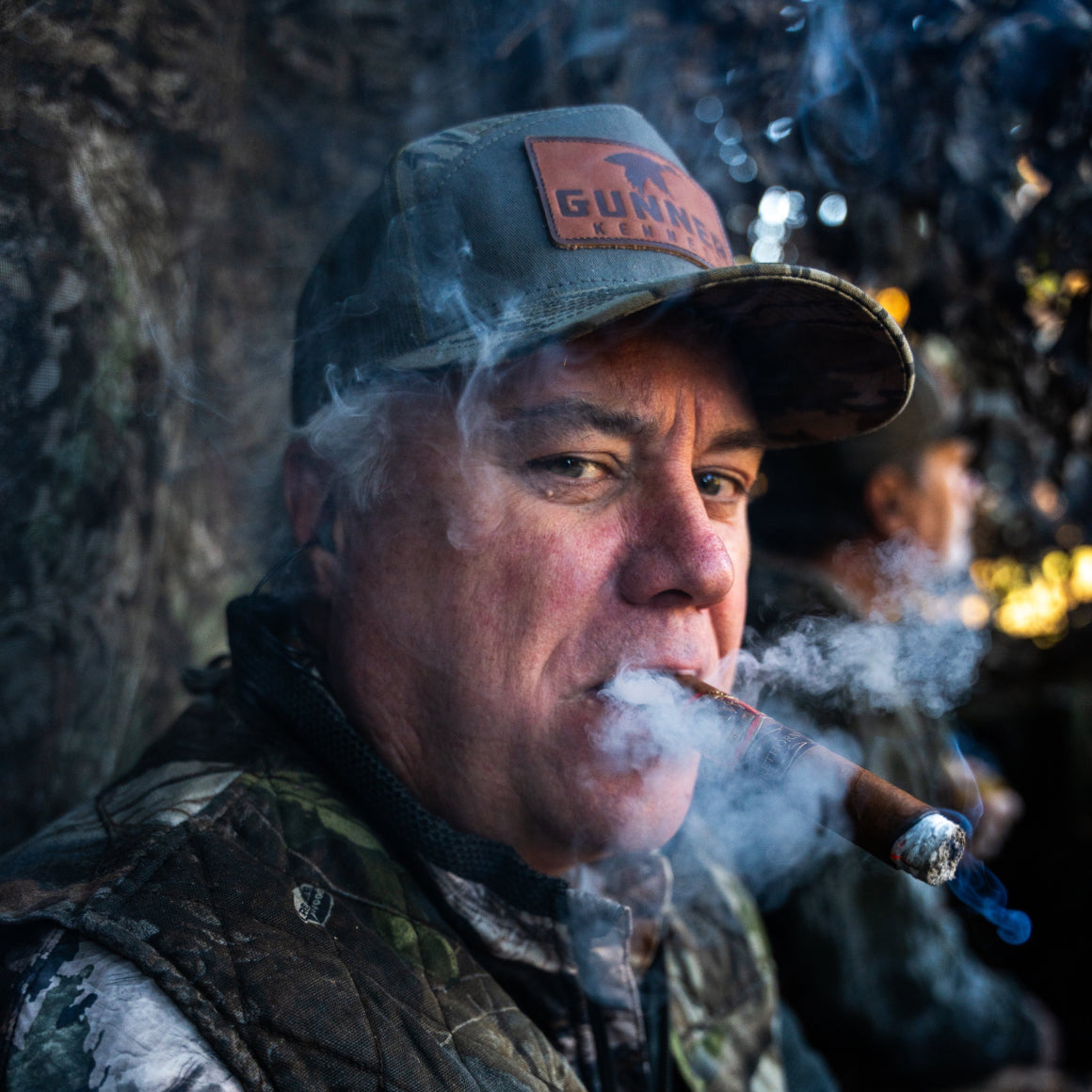 A man in camouflage and a GUNNER BOTTOMLAND HAT smokes a cigar outdoors, exhaling smoke. Another person is blurred in the background among the trees.