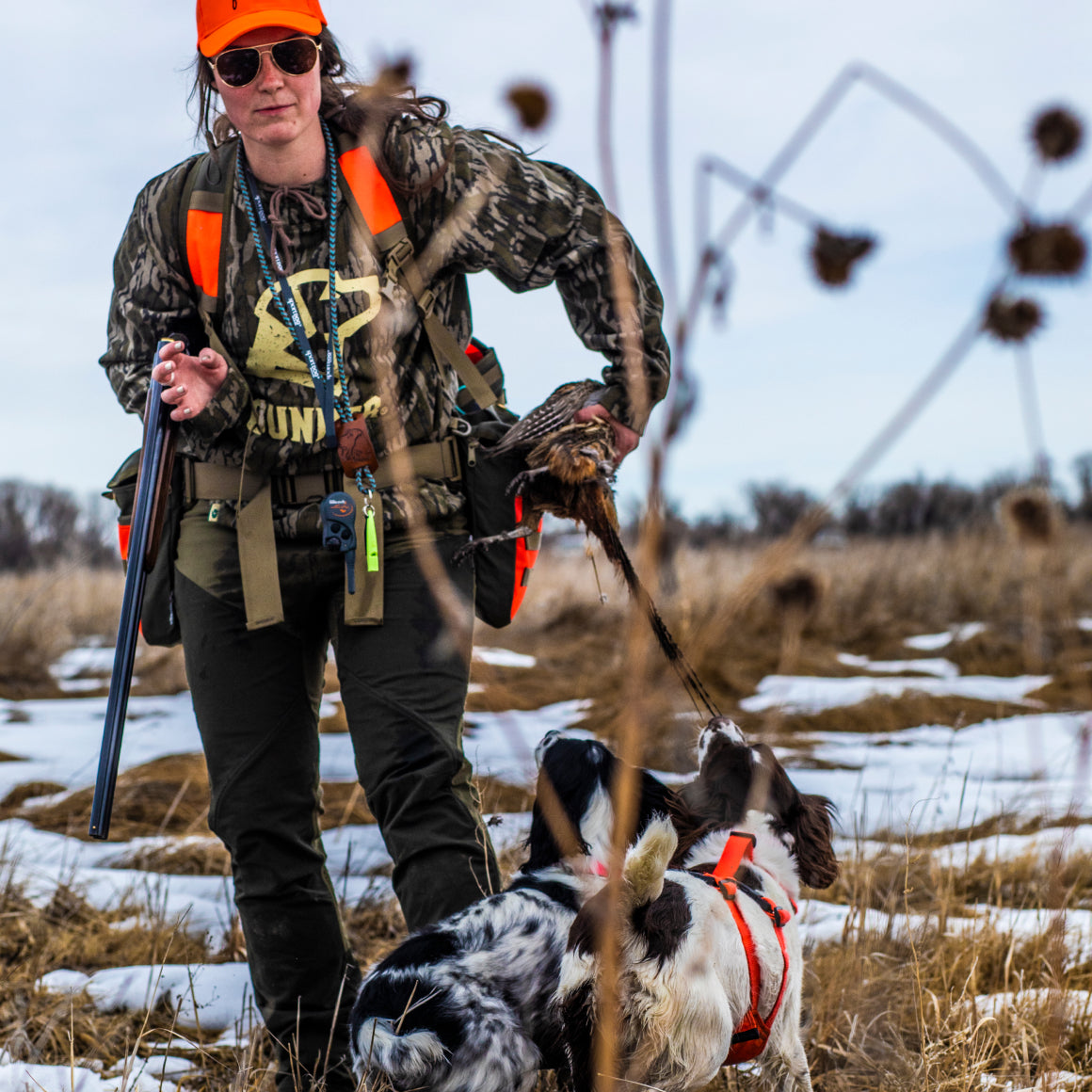 Wearing the GUNNER BOTTOMLAND HOODIE and an orange hat, a hunter holds a shotgun and a game bird as two dogs look up at her in a snowy grassy field.