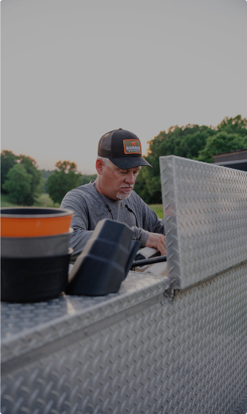 A man wearing a cap and grey shirt works at the back of a metal truck with a diamond plate pattern, outdoors near green trees under a clear sky. Plastic buckets and a dog crate are stacked on the truck.