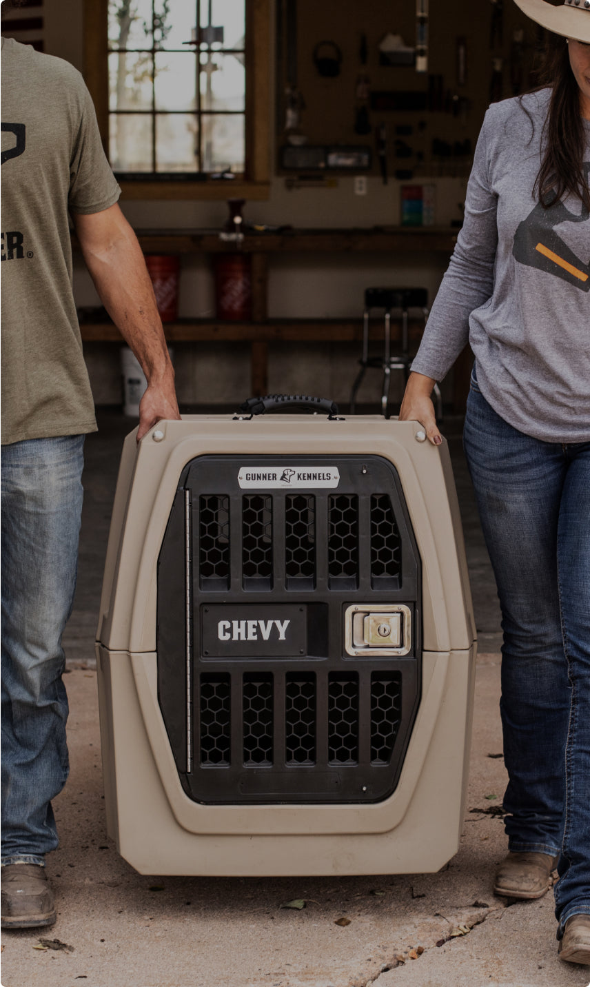Two people stand on either side of a large beige and black dog crate labeled CHEVY in a garage or workshop setting, surrounded by outdoor gear. Only their torsos and arms are visible.