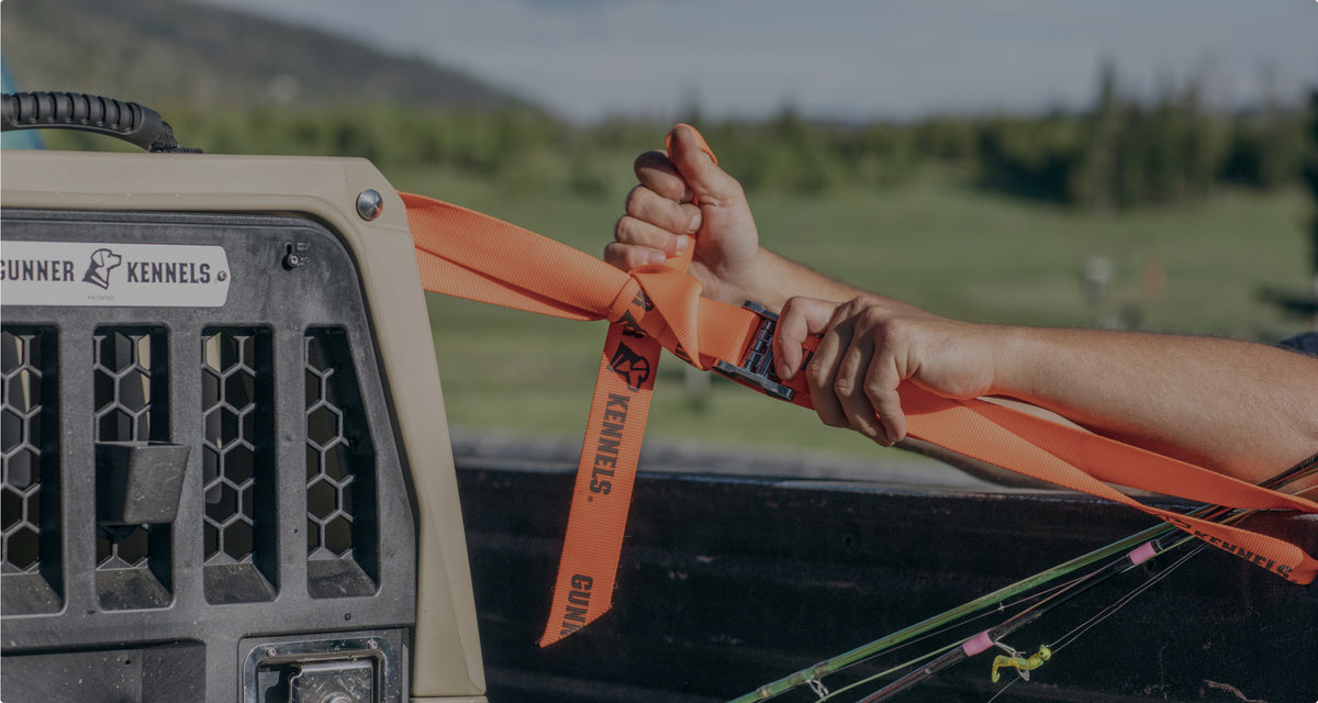 A person secures an orange strap labeled GUNNER KENNELS to a tan dog kennel in the bed of a truck, surrounded by green fields and hills—ideal outdoor gear for any working dog.