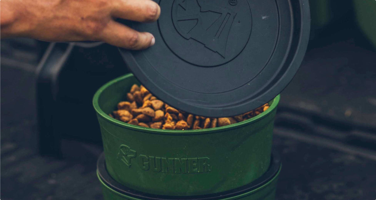 A hand lifts the lid off a green container filled with dry dog food. The container, perfect for storing outdoor gear or fitting inside a dog kennel, has the brand name GUNNER embossed on its side and sits on a dark surface.