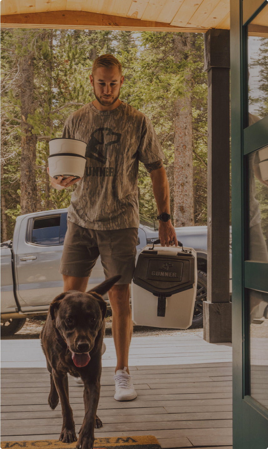A man in shorts and a camouflage shirt carries a white cooler and outdoor gear while stepping onto a porch, followed by a happy chocolate Labrador. A silver pickup truck is parked in the background among tall trees.