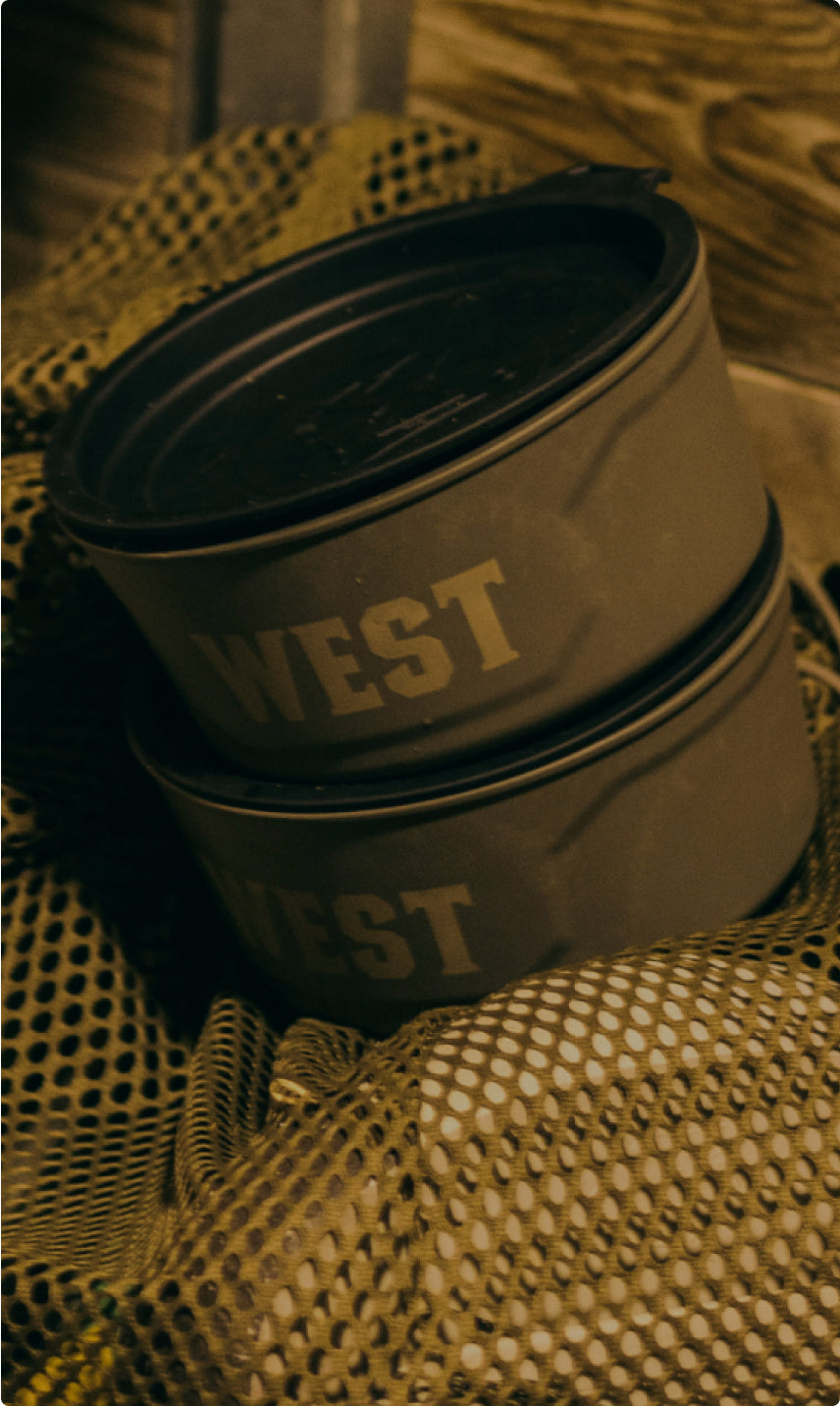 Two stacked, round olive-green containers with the word WEST printed on the side, placed on a mesh netting near a dog crate and a wooden surface in the background.