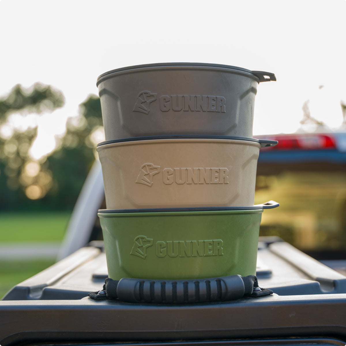 Three stackable dog bowls in gray, tan, and green—embossed with the Gunner logo—are placed on the back of a vehicle outdoors, blending seamlessly with your kennel setup and essential outdoor gear. Trees and sunlight complete the perfect working dog scene.
