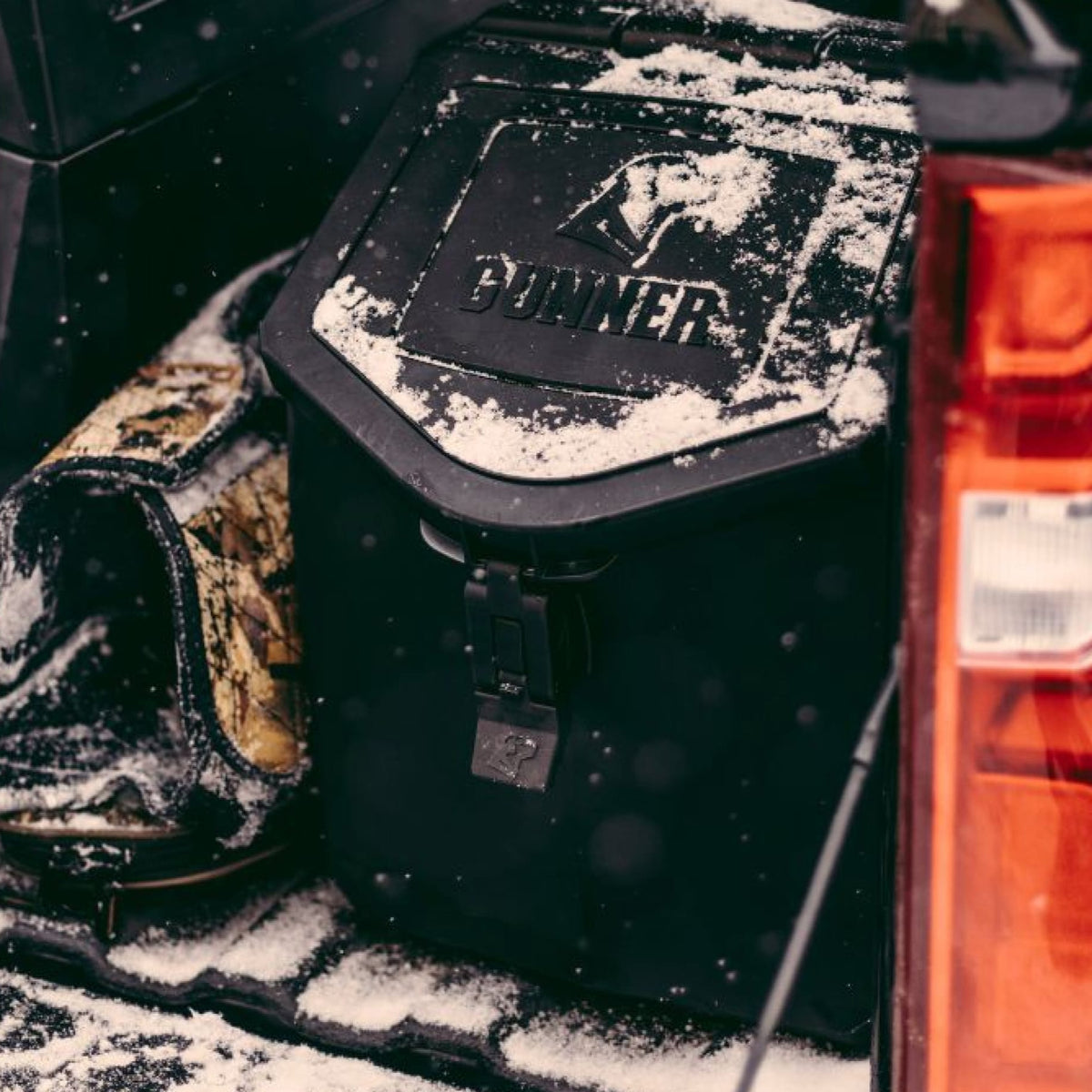 A black GUNNER FOOD CRATE by GUNNER, dusted with snow, rests in a vehicle beside a camouflage item and an odor-proof dog food container near a red tail light—ideal for winter adventures or travel with your pet.