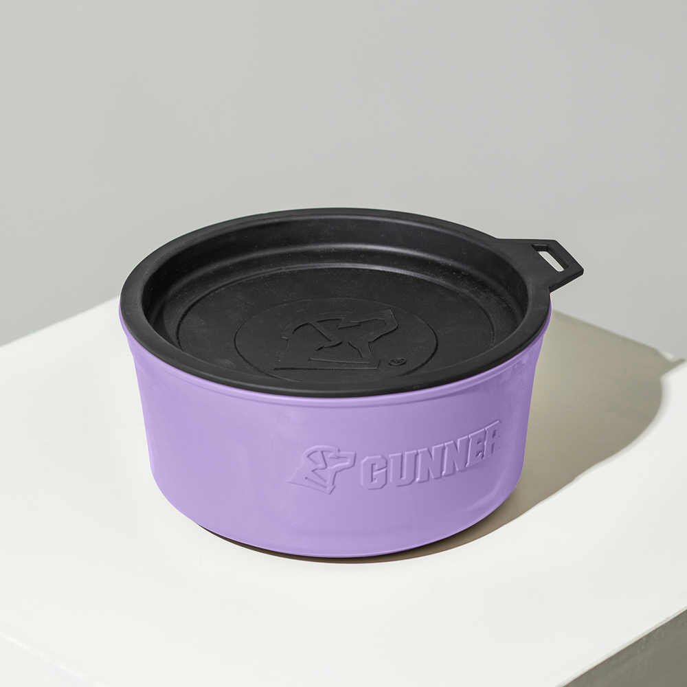 The limited-edition GUNNER DOG BOWL - LAVENDER by GUNNER features a round shape, black lid, dog logo, and Color Fetch design. It’s shown on a white surface with a plain background.
