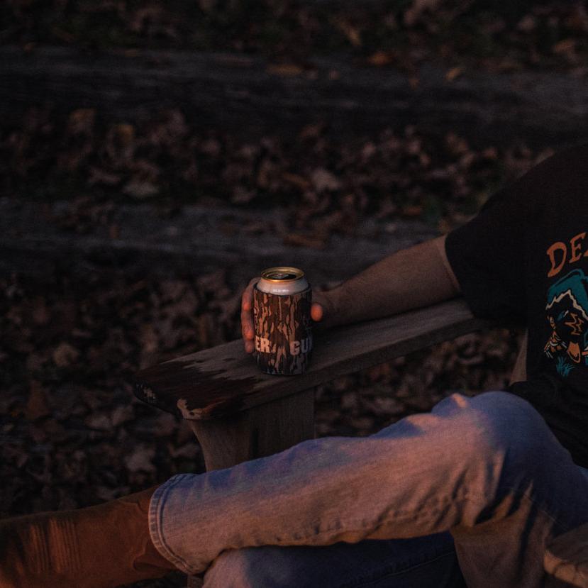 Wearing jeans and a dark graphic tee, someone sits on a wooden chair outdoors, holding a can with the GUNNER KOOZIE - Bottomland by GUNNER. Fallen leaves cover the ground, setting an autumn evening mood.