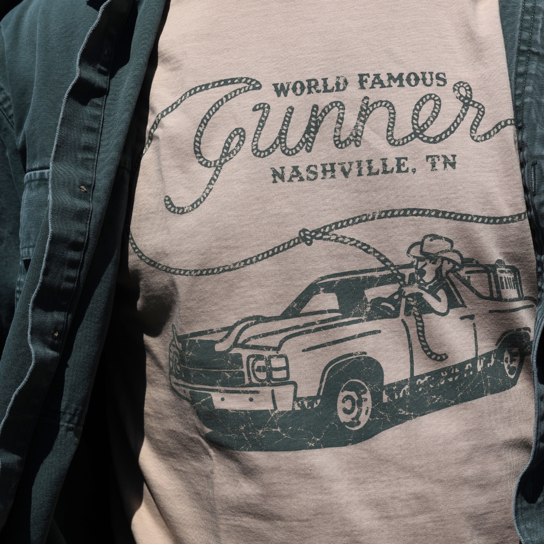 A person wears GUNNER’s tan WORLD FAMOUS TEE featuring World Famous Gunner Nashville, TN and a cowboy driving a vintage pickup, layered under an open dark green shirt for Southern style.