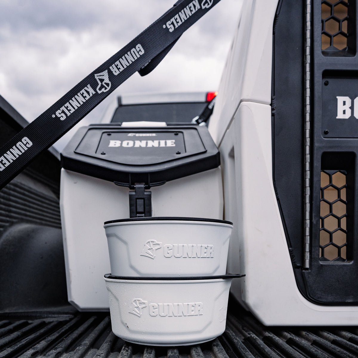 Two white GUNNER DOG BOWL - GUNNER YARD bowls from GUNNER are stacked in front of a pet travel crate labeled BONNIE in a truck bed, with a black GUNNER leash nearby.