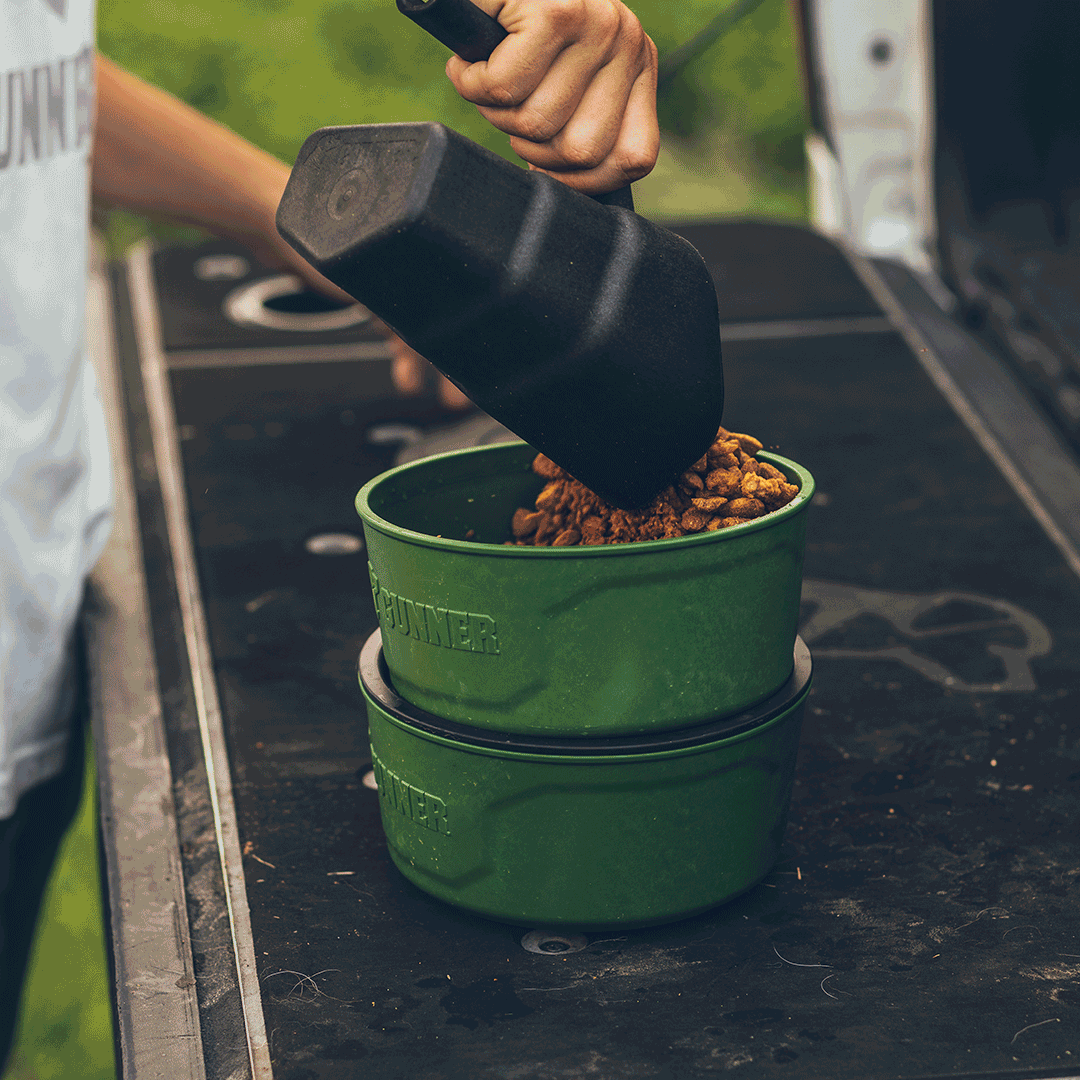 A person scoops kibble into a green pet bowl outdoors, using a black scoop—perfect for a working dog. Another empty green bowl is stacked underneath on a black surface.
