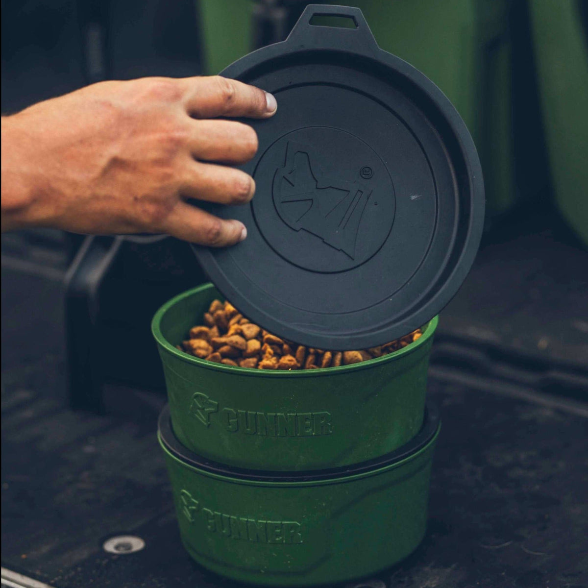 A hand lifts the lid off a GUNNER DOG BOWL - DUCK GREEN filled with dry pet food, perfect for refills; another empty GUNNER bowl is stacked beneath it.