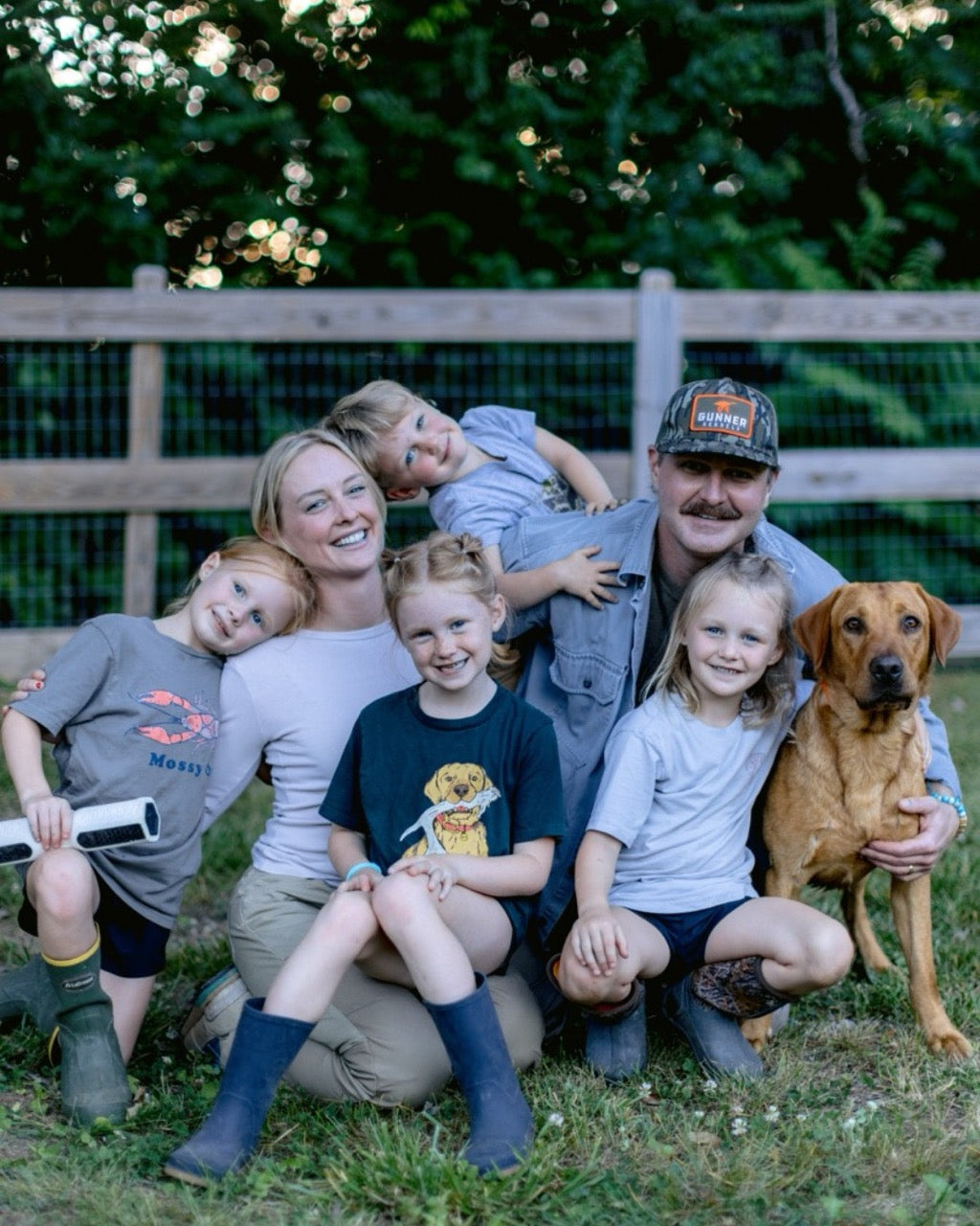 A smiling family of two adults, four young children, and a brown working dog pose together outdoors in front of a wooden fence and trees. The casually dressed group appears happy with their loyal companion from the local dog kennel.