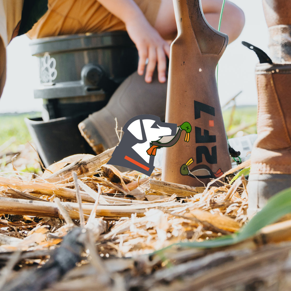 A close-up of boots and outdoor gear, including a rifle lying on dry corn stalks outdoors, with a bucket and a persons hand visible in the background. A dog kennel logo sticker is on the rifle.