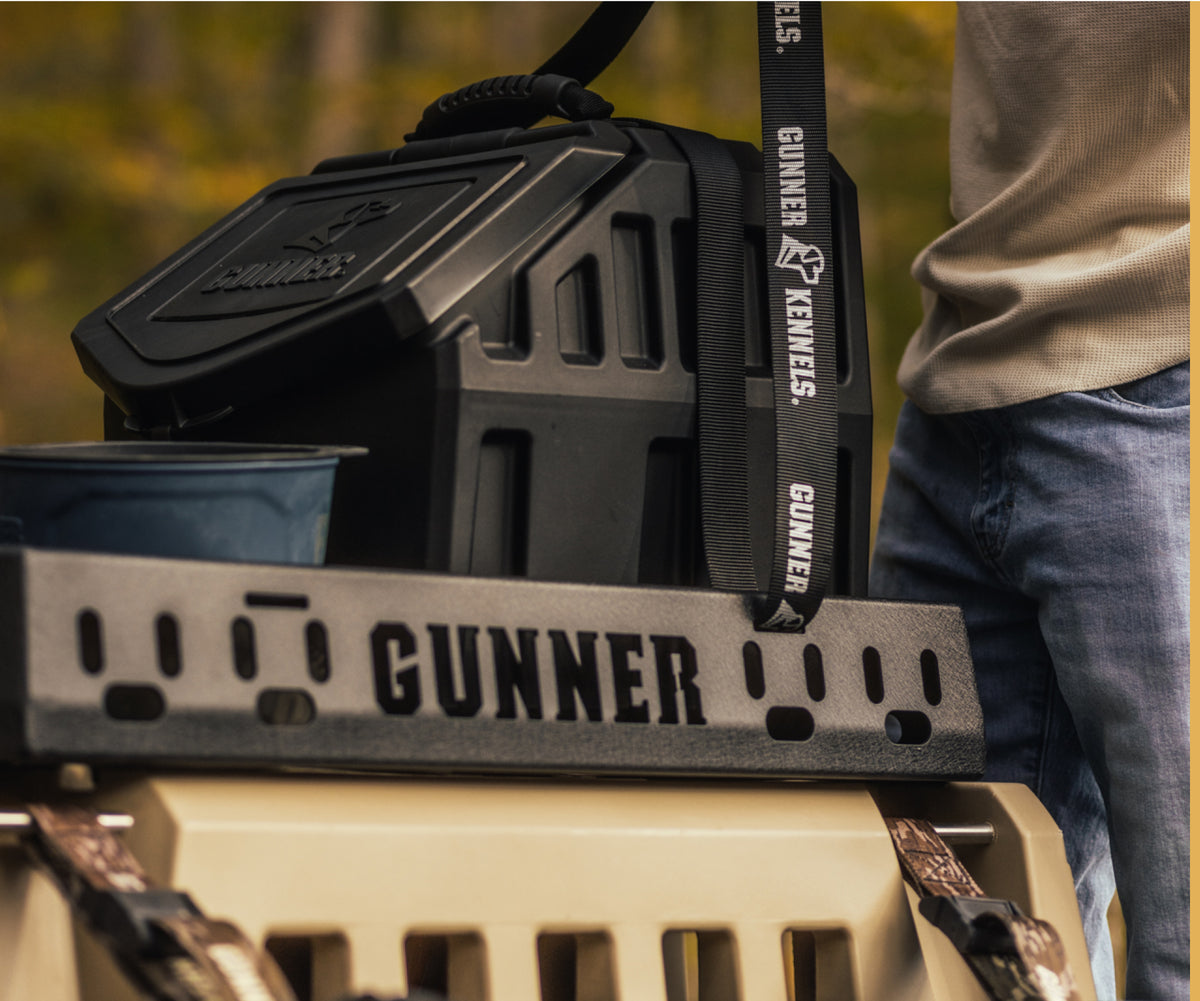 A person stands next to a sturdy, black GUNNER dog kennel and a matching crate with a strap labeled GUNNER KENNELS outdoors, surrounded by trees blurred in the background—a perfect setup for outdoor gear and working dog adventures.