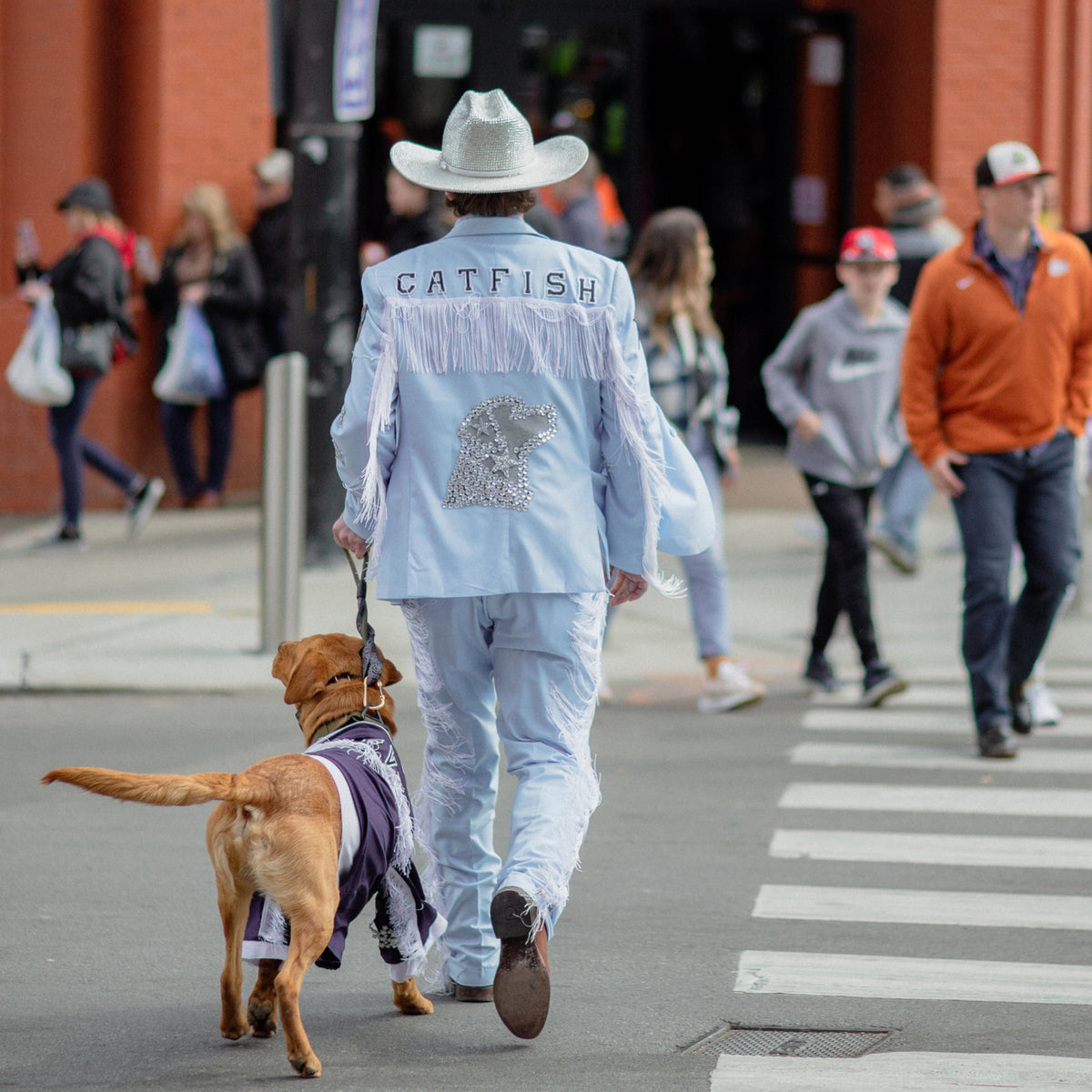 A person in a light blue fringed suit with CATFISH and a sequin fish on the back walks a golden retriever working dog in a vest across a city crosswalk, with pedestrians and a brick building in the background.