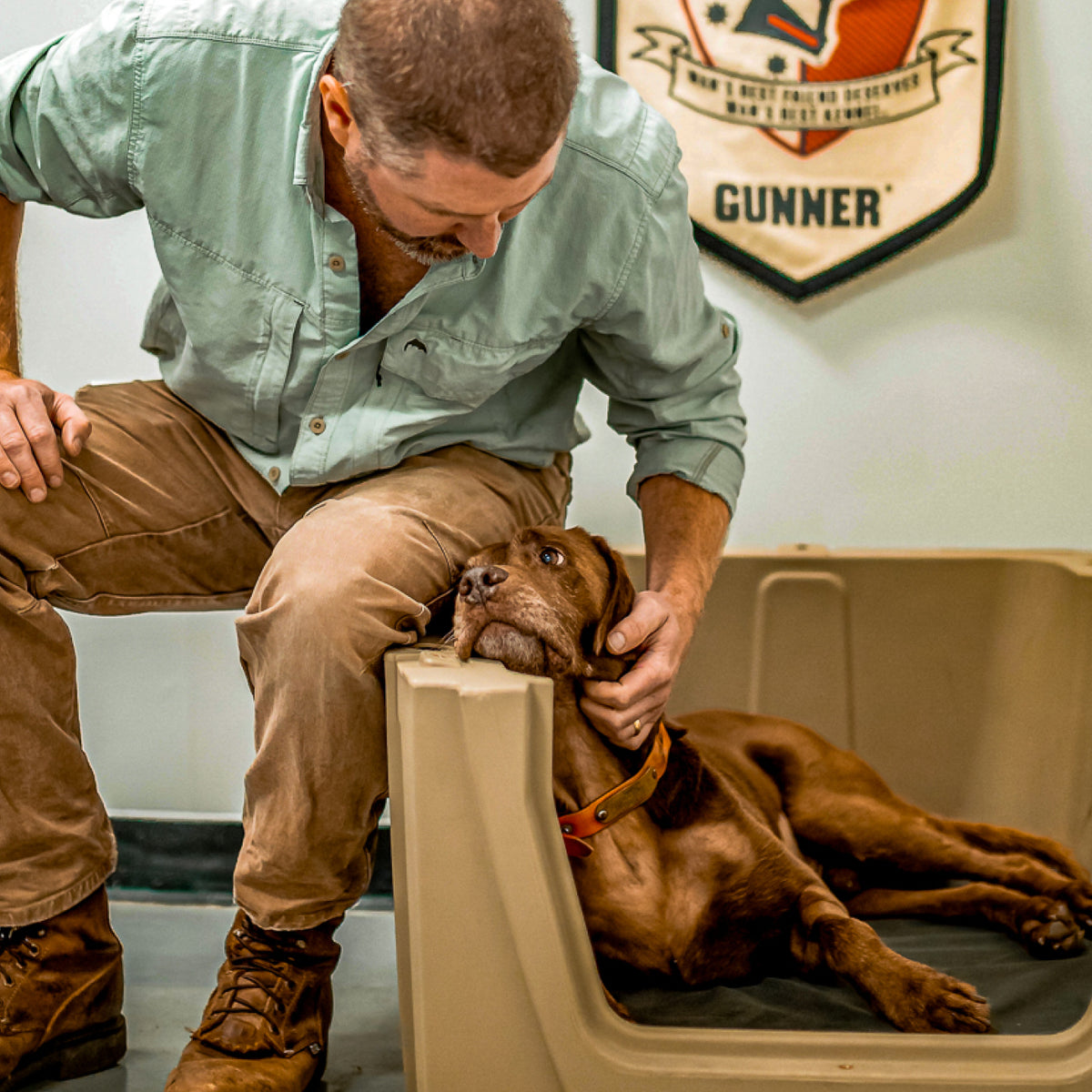 A man kneels beside a brown dog lying in a plastic dog kennel, gently holding the dog’s head. A badge with the name Gunner hangs on the wall in the background.