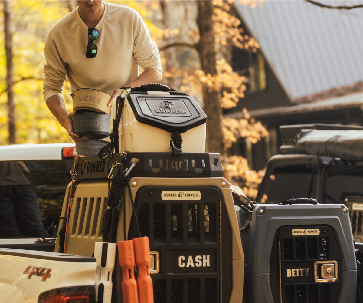 A person loads labeled dog kennels, including ones marked CASH and BETTY, along with outdoor gear, into the back of a pickup truck, with trees and a cabin visible in the background.