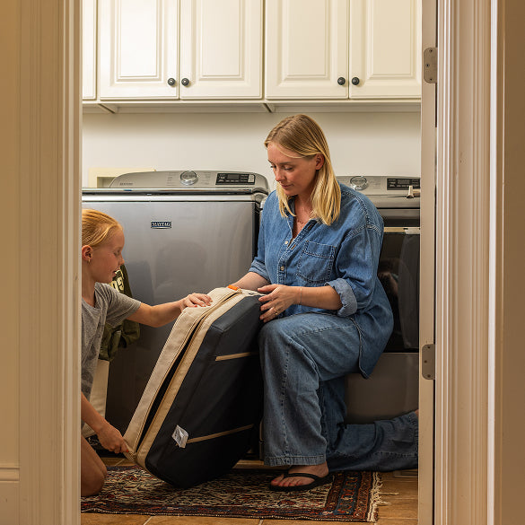 A woman in denim sits on the floor of a laundry room next to a washing machine, helping a child with light hair hold a large black and beige laundry bag. A dog crate rests nearby, and white cabinets are visible above the machines.