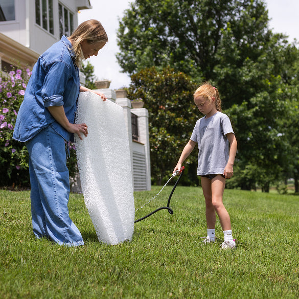 An adult and a child stand on a grassy lawn. The adult holds a large sheet of white bubble wrap, while the child uses a garden hose to spray water onto it. Nearby, a dog kennel and some outdoor gear are visible among the trees and house in the background.
