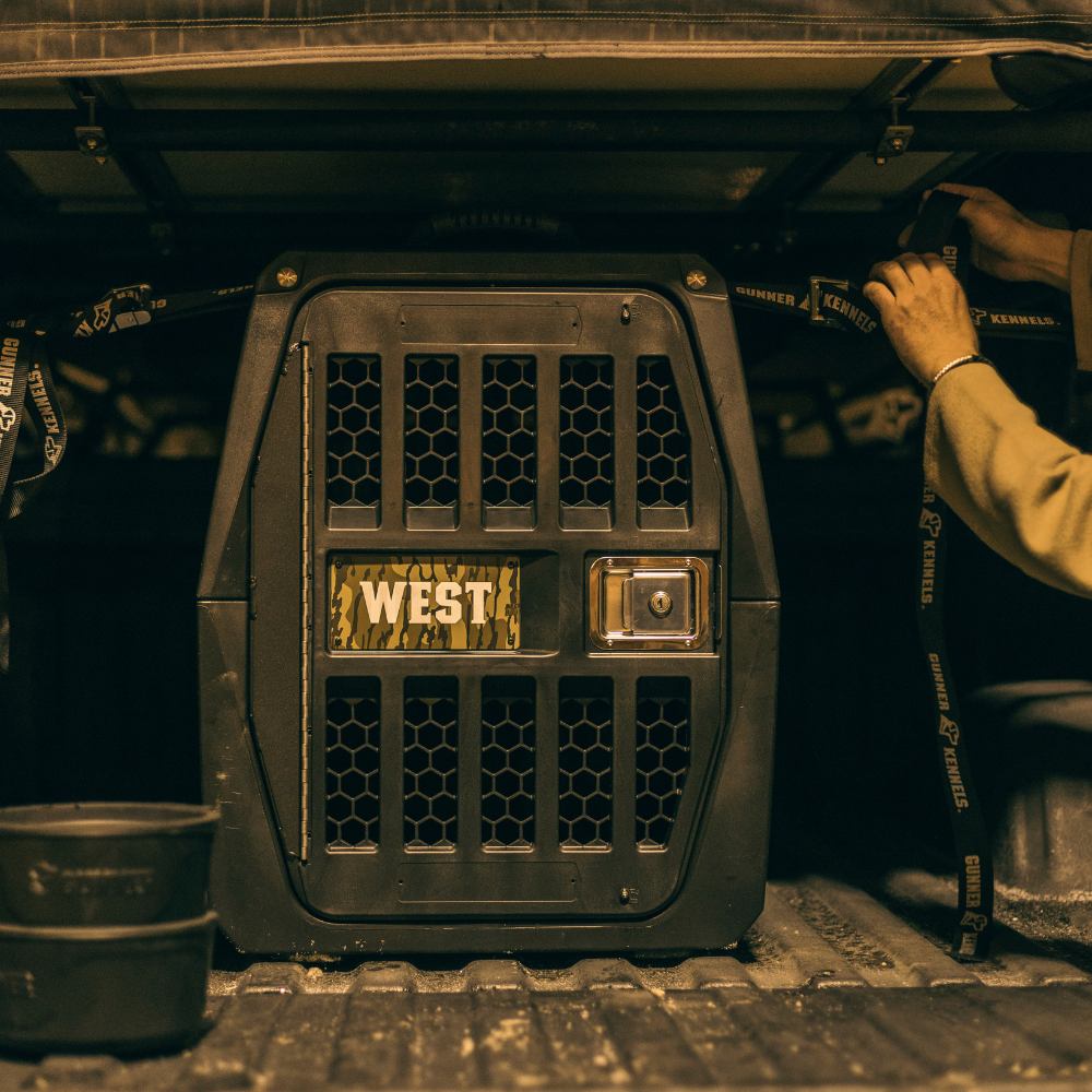 A person secures the GUNNER G1™ KENNEL - BLACK with camouflage interior inside a vehicle using straps. The kennel sits on a textured surface, with two stacked bowls positioned nearby.