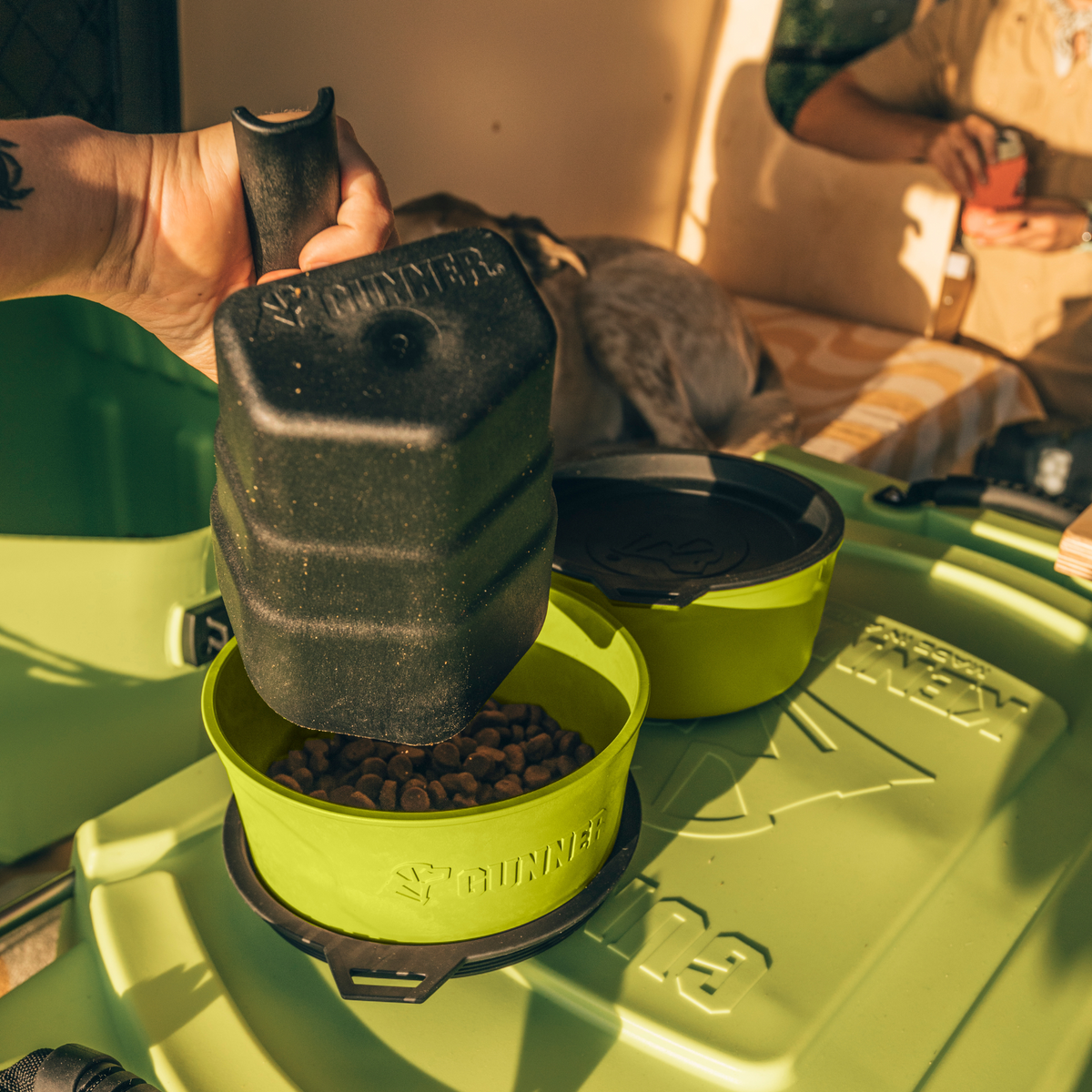 A hand pours dry food from a limited-edition Color Fetch black container into the GUNNER DOG BOWL - SULPHUR SPRINGS, set on a green storage box as a dog relaxes nearby in warm light.