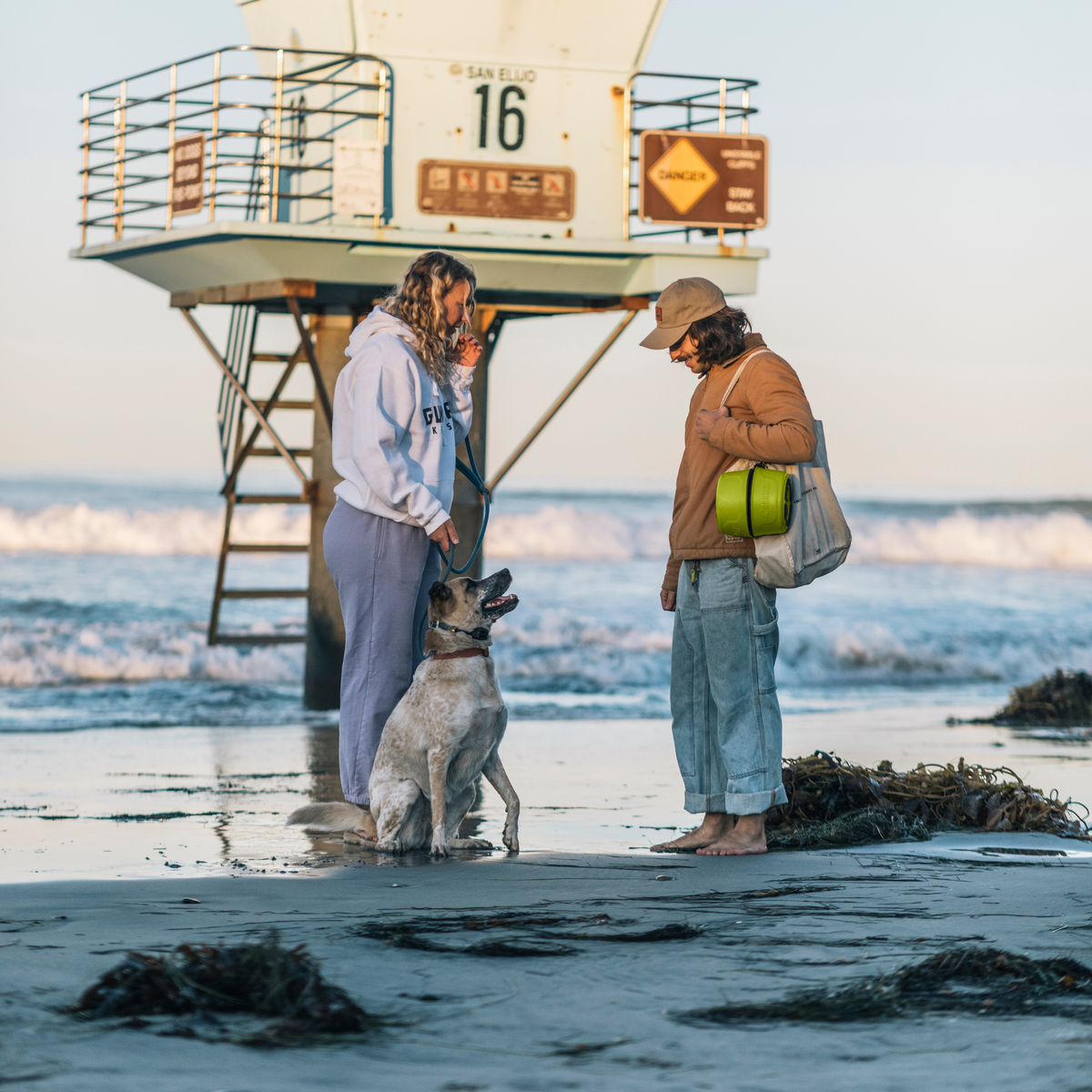 Two people stand barefoot on the beach by a lifeguard tower, chatting while their dog sits between them with a GUNNER DOG BOWL - SULPHUR SPRINGS from GUNNER nearby. Seaweed covers the wet sand and ocean waves roll in for a perfect coastal moment.