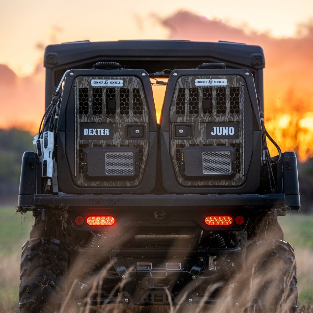 A rugged off-road vehicle at sunset features two GUNNER G1™ KENNEL - MOSSY OAK BOTTOMLAND BLACK crates, labeled “Dexter” and “Juno,” mounted on the back. Tall grass surrounds the vehicle, with red tail lights glowing in the dusk.
