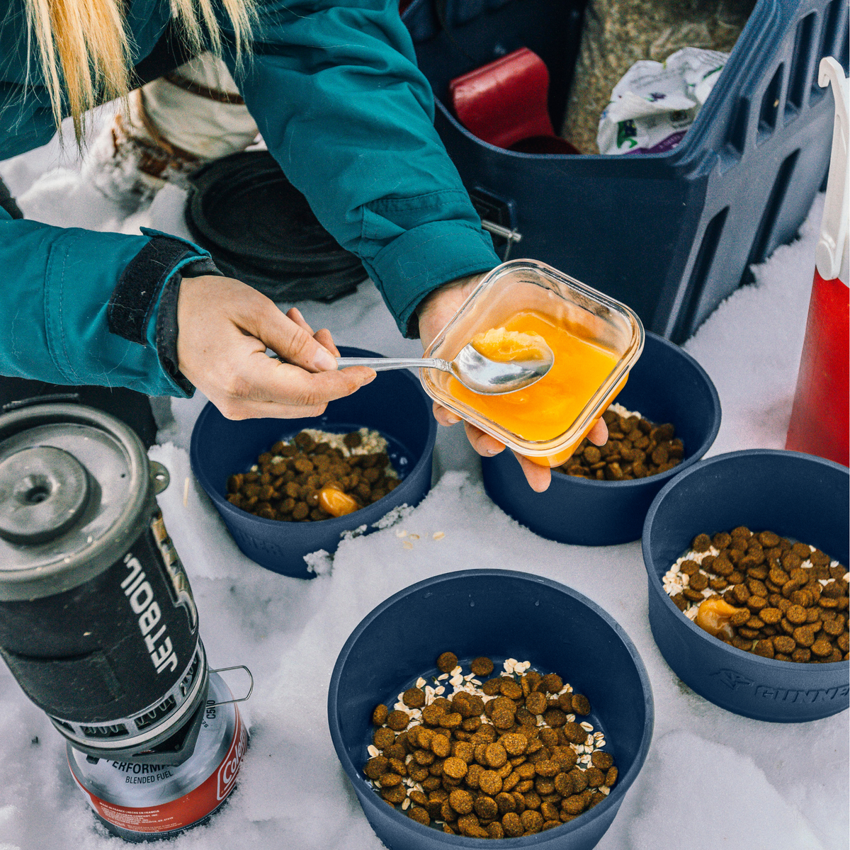 Wearing a limited-edition teal jacket, a person adds orange liquid to pet food in GUNNER DOG BOWL - NAVY by GUNNER, set on snow with camping gear and a navy portable stove close by.