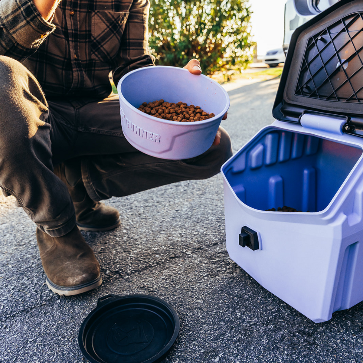 Wearing boots and a plaid shirt, a person scoops dry pet food from a GUNNER DOG BOWL - LAVENDER by GUNNER on a paved outdoor surface, with sunlight and greenery in the background.