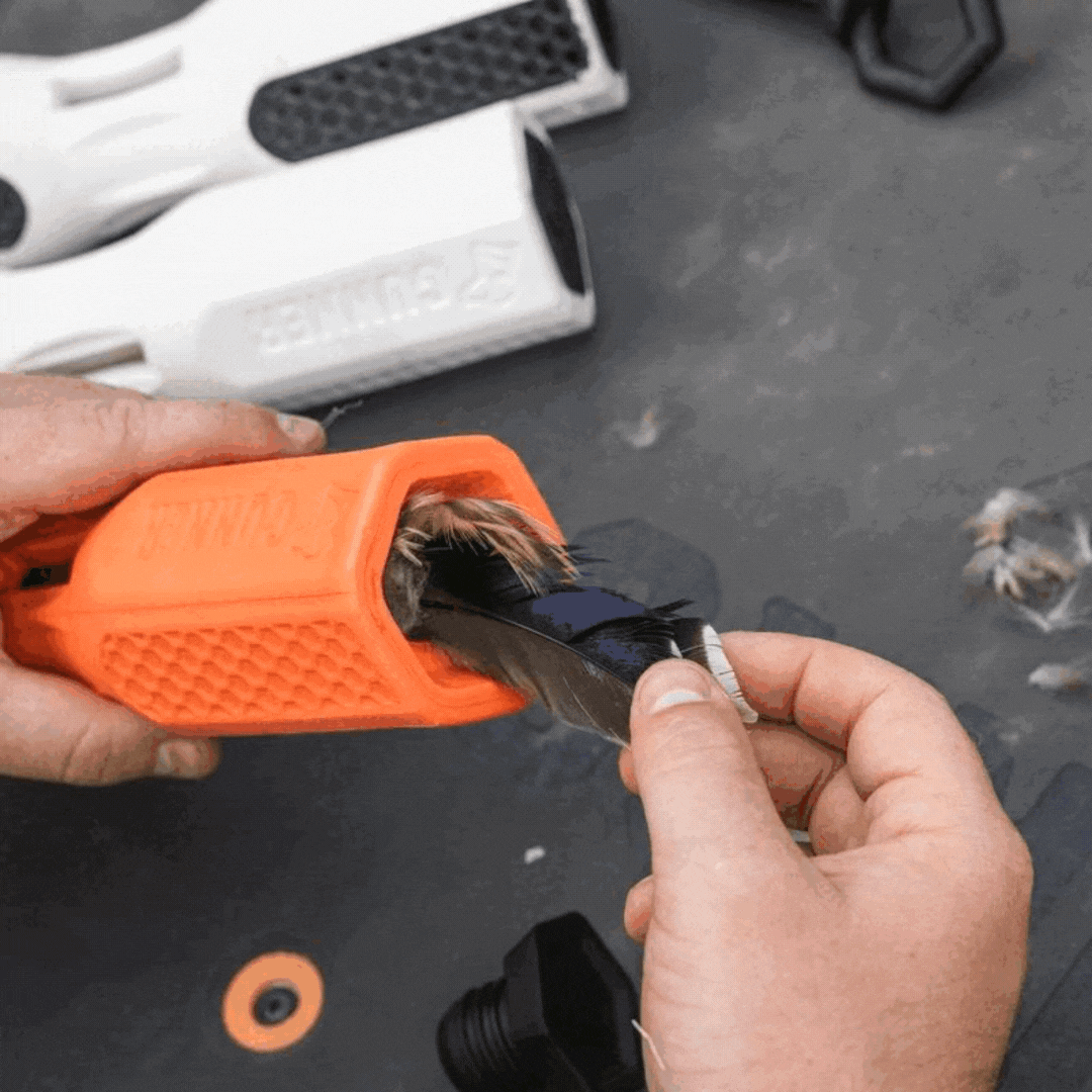 Close-up of hands using the orange GUNNER BUMPER by GUNNER to remove feathers from a bird wing, with additional tools and feathers visible on a gray surface in the background.