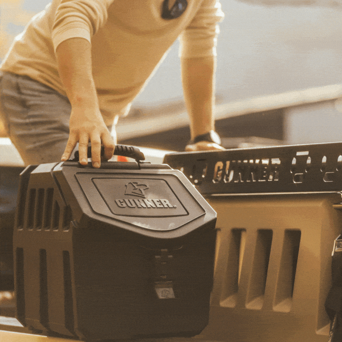 A person wearing a tan sweater and sunglasses places a sturdy black GUNNER dog crate onto the back of a pickup truck in sunlight. The kennel, known for its tough outdoor gear design, features a handle on top.