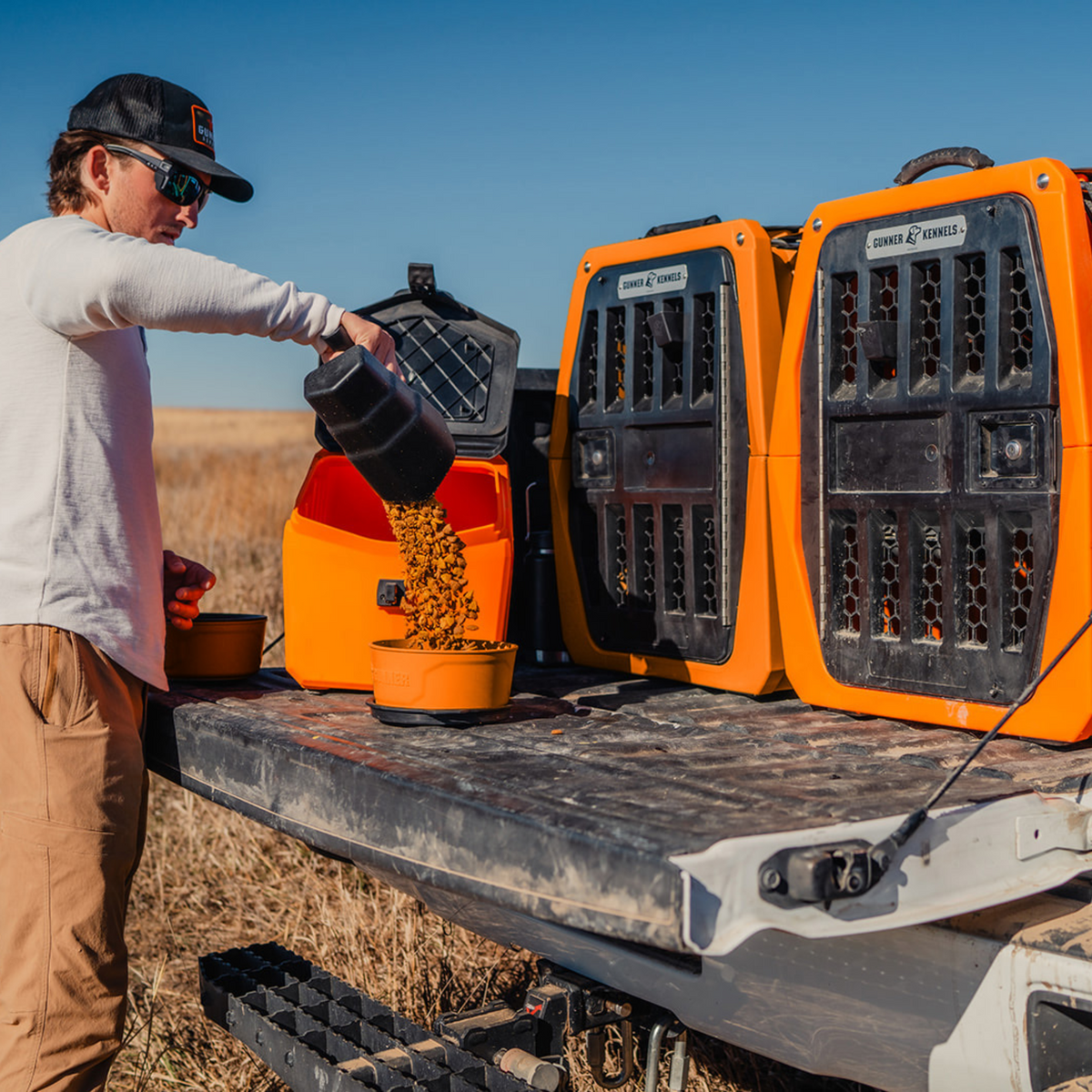 Wearing outdoor gear, a person pours dog food from a GUNNER FOOD CRATE - BLAZE ORANGE into a bowl on a pickup tailgate, with two bright orange GUNNER crates nearby in a grassy field beneath a clear blue sky.