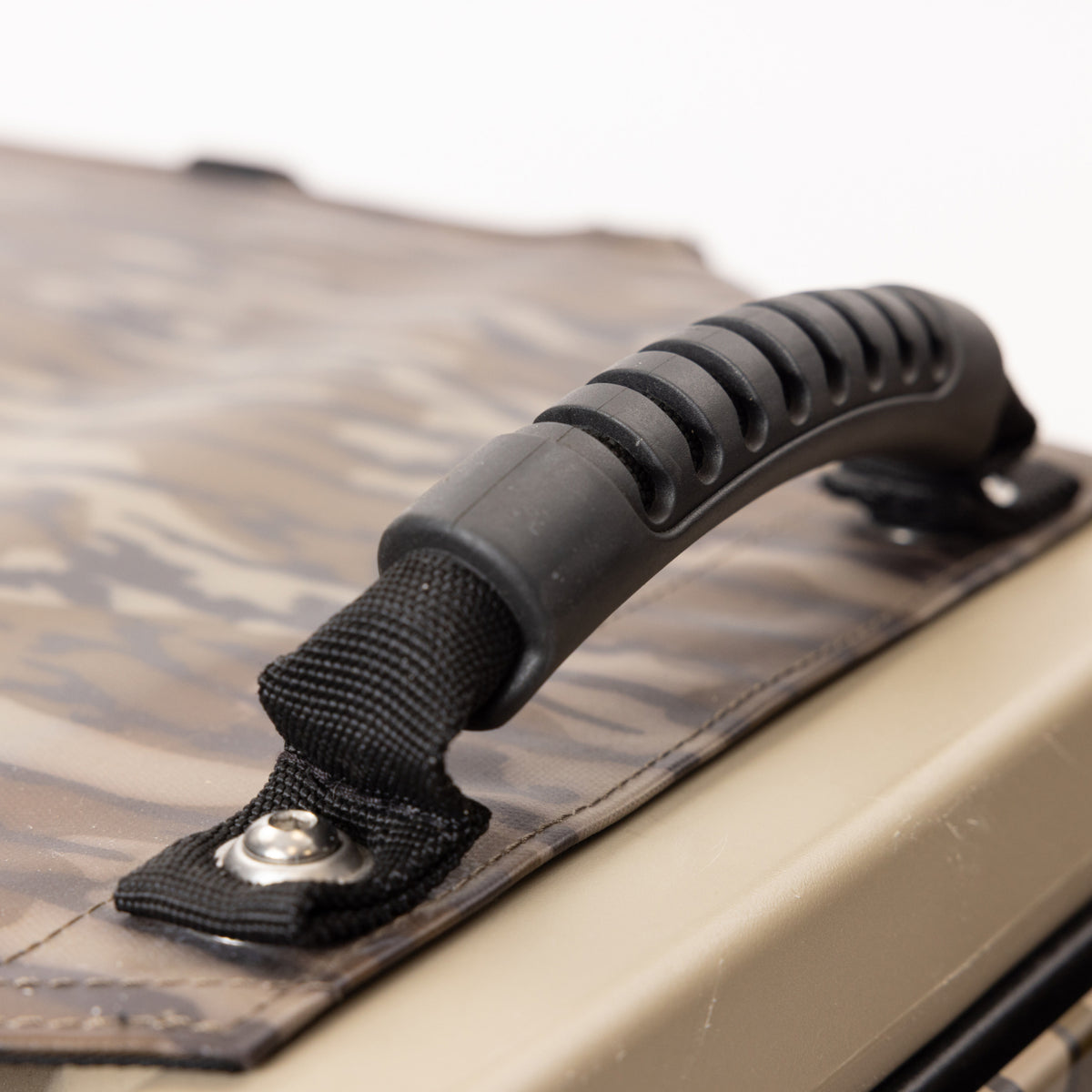 Close-up of a black plastic and fabric handle attached to a GUNNER ALL-WEATHER KIT 2.0 MOSSY OAK BOTTOMLAND bag, featuring a camouflage pattern, secured with metal rivets, and designed for enhanced UV protection.