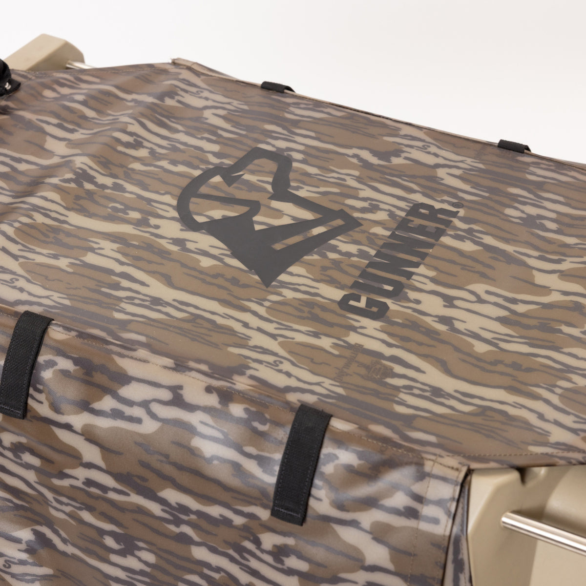 A close-up of the GUNNER ALL-WEATHER KIT 2.0 MOSSY OAK BOTTOMLAND, a camouflage-patterned waterproof kennel cover featuring the GUNNER logo and stylized dog head, securely attached to a beige surface with black straps for added security and UV protection.