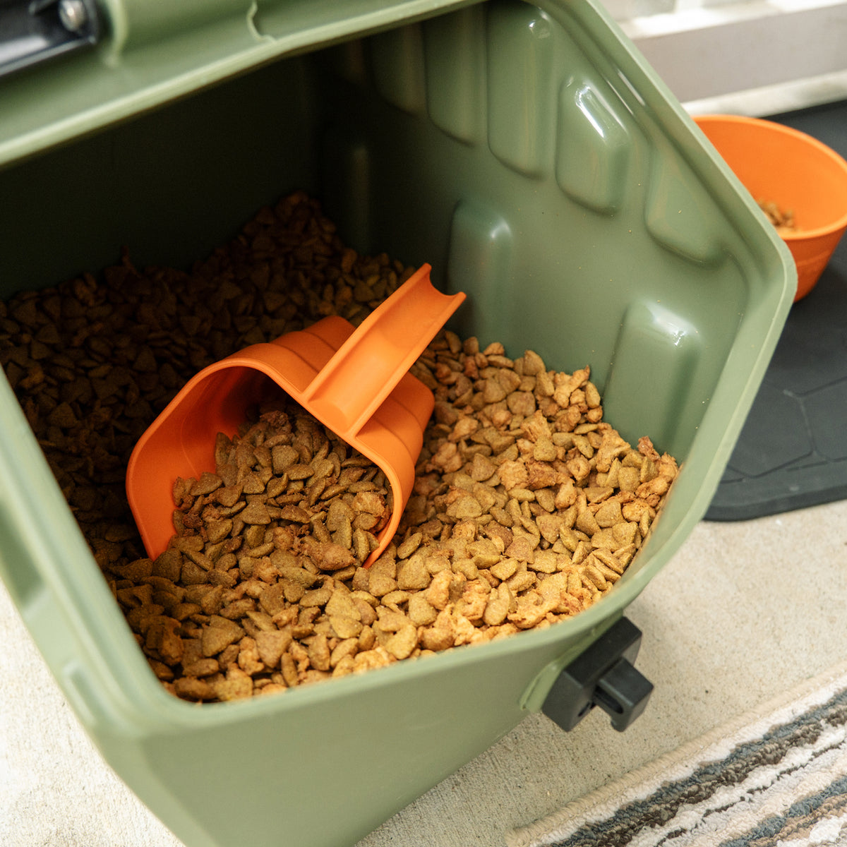 A GUNNER FOOD SCOOP BLAZE ORANGE filled with dry kibble inside an open olive green storage bin.