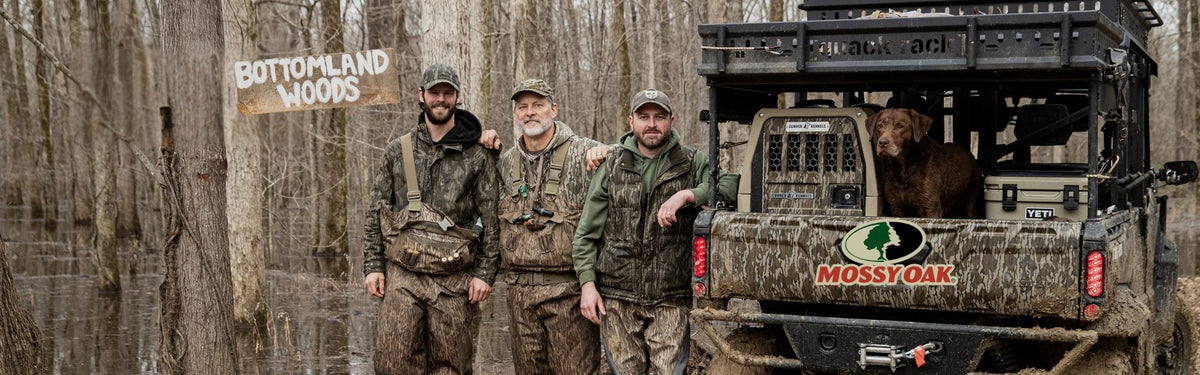 Four men in camo gear stand by an off-road vehicle with a hunting dog in a kennel in the back. They’re in a flooded, leafless forest next to a Bottomland Woods sign. The vehicle is equipped with outdoor gear and features a Mossy Oak logo.