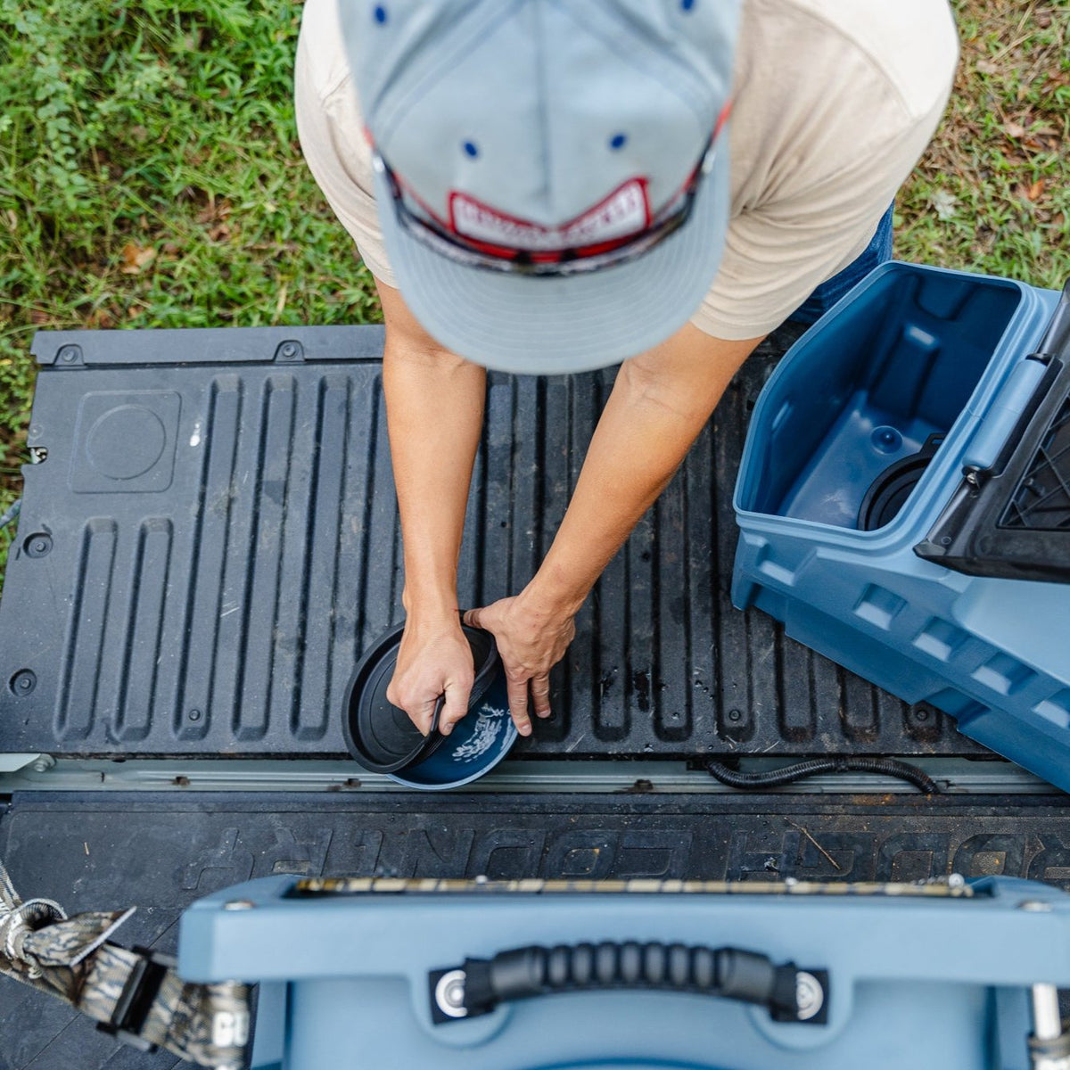 A person is seen from above using a GUNNER DOG BOWL - Ducks Unlimited Blue portable bowl by GUNNER to fill water for their dog on a truck tailgate near grass—ideal after exploring waterfowl habitats.