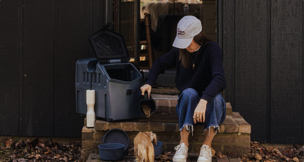 A woman in a white cap and dark sweater sits on brick steps, scooping food for her small tan hunting dog outside a dark wooden building, with bowls and pet supplies nearby.