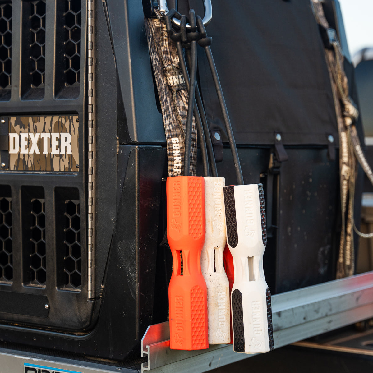 Three bone-shaped GUNNER Bumpers—a Bumper Bundle with orange, white, and black—hang by cords from a vehicle crate labeled Dexter, which is secured with camo and black webbing straps for safe travel.
