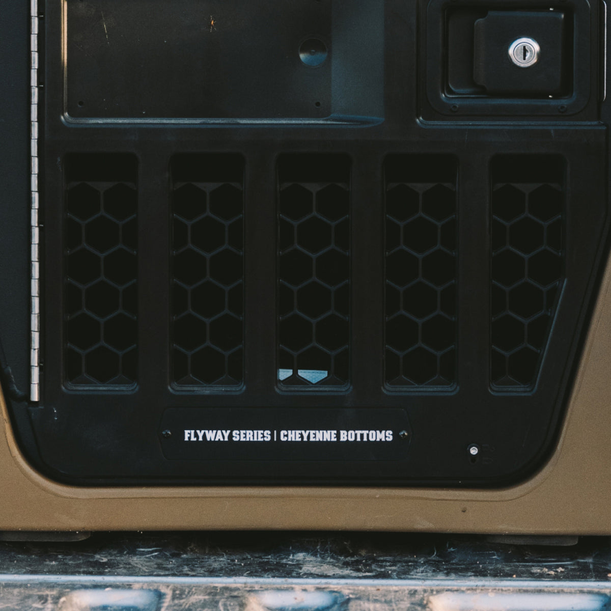 A tan and black Kennell dog crate sits in the back of a pickup truck, surrounded by hunting gear including a hanging deer head and other equipment. A door tag reads Flyway Series | Cheyenne Bottoms