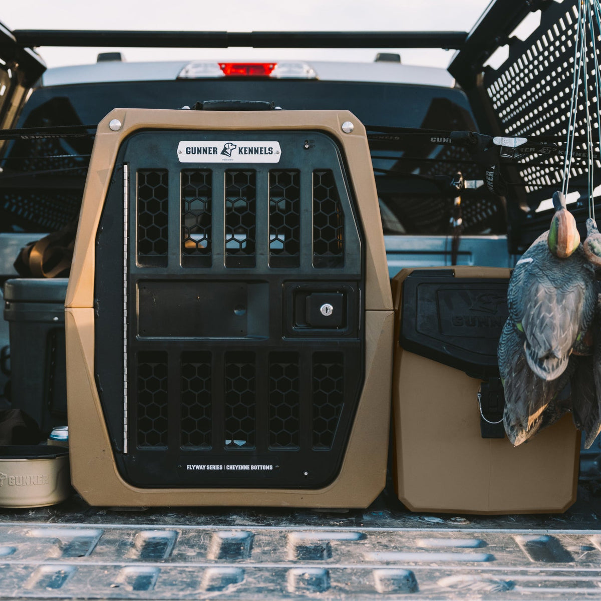 A tan and black GUNNER G1™ KENNEL – Cheyenne Bottoms sits in the bed of a truck, surrounded by gear and two hanging ducks—perfect for your next adventure through the Central Flyway or a morning at Cheyenne Bottoms. The kennel features ventilation holes, a secure door, and proudly displays the GUNNER label on the front.