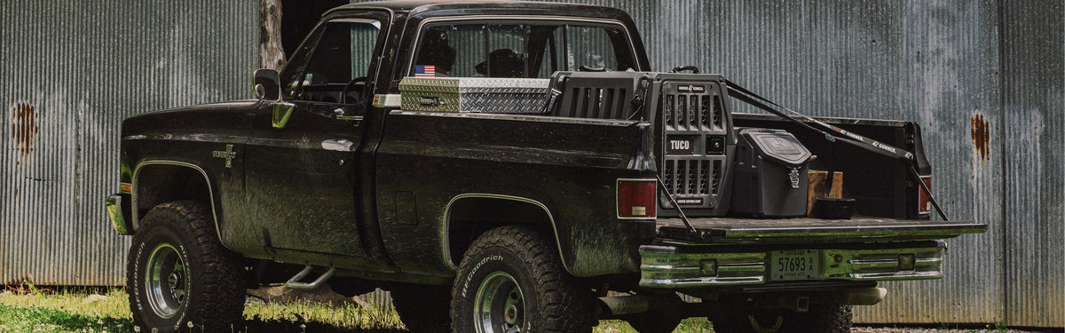 A black vintage pickup truck with rugged tires is parked on grass near a corrugated metal building, with outdoor gear and a toolbox in its bed.